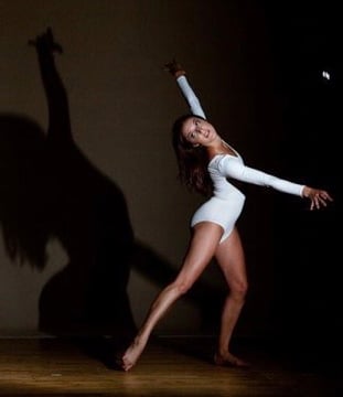 A dancer in a white leotard with a spotlight on her and her shadow on the wall.