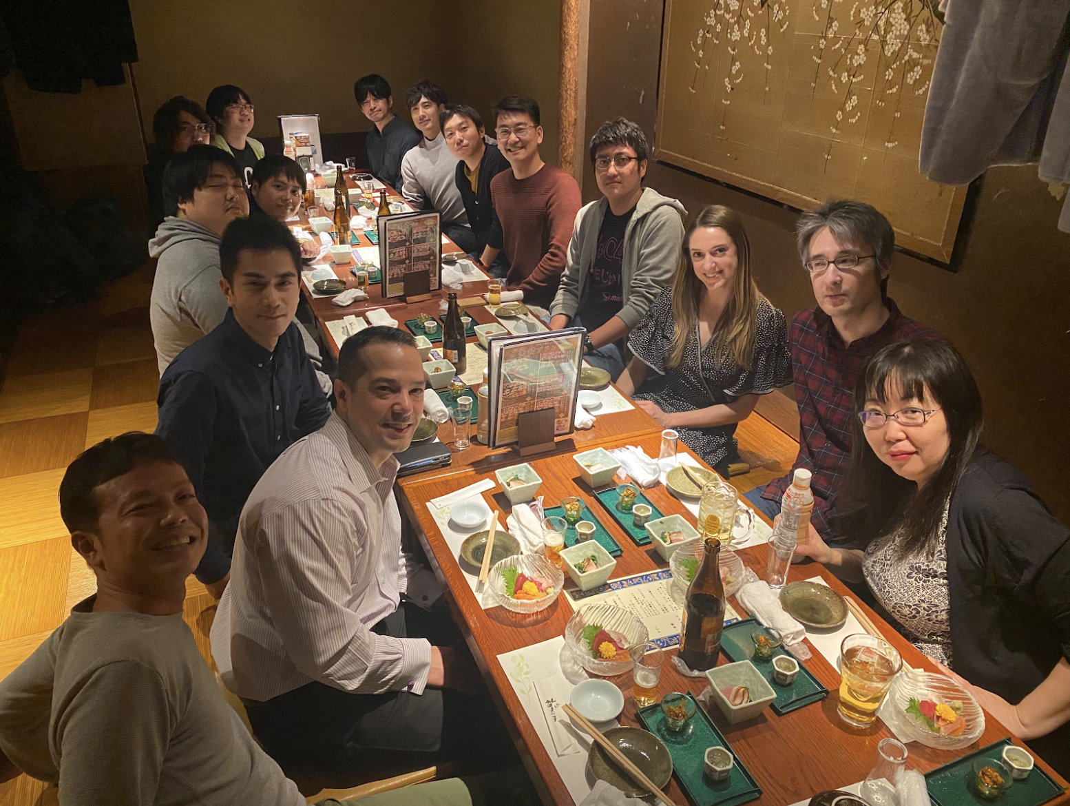 A large group of people seated at a very long table having dinner. They are all smiling and looking at the camera.