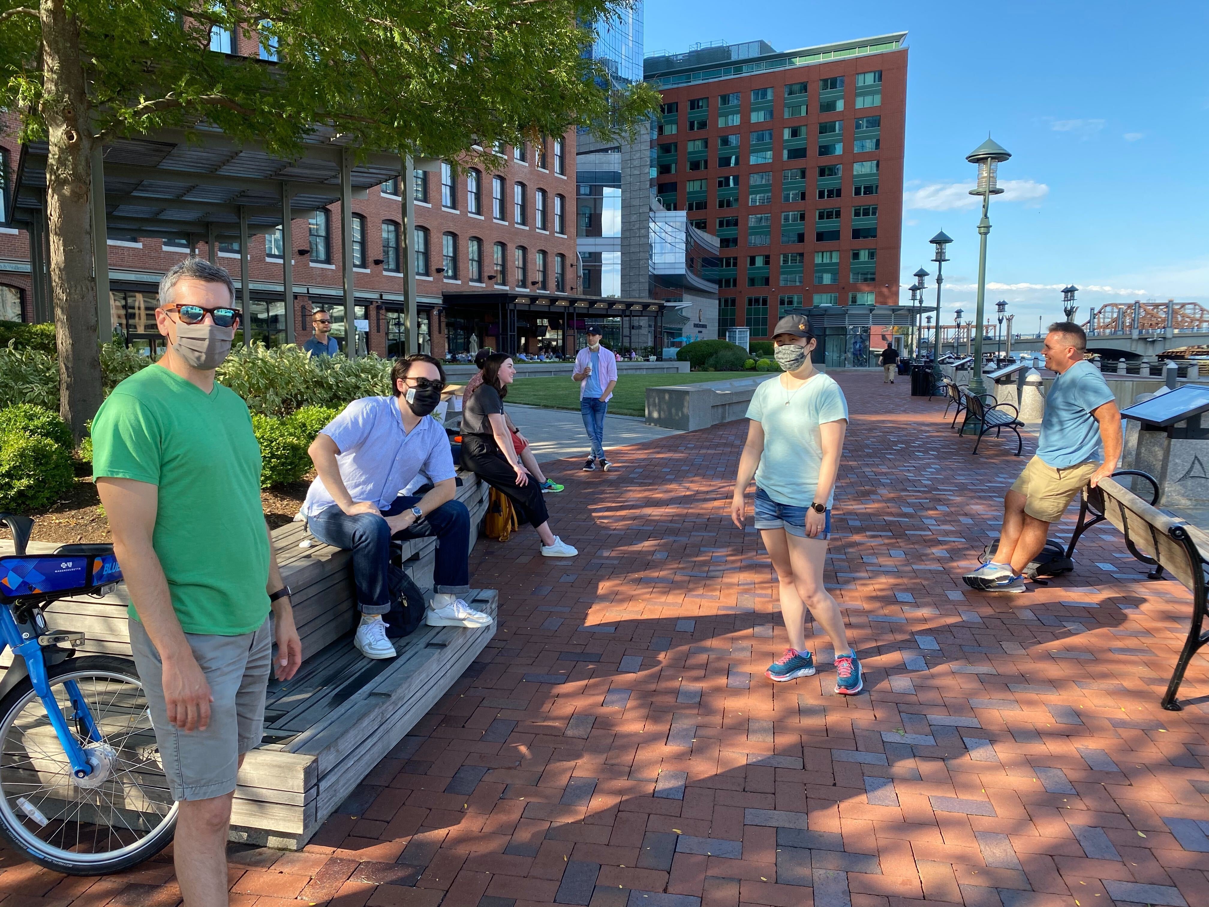 Several people wearing face masks, standing and walking around a brick walkway in front of some tall buildings.