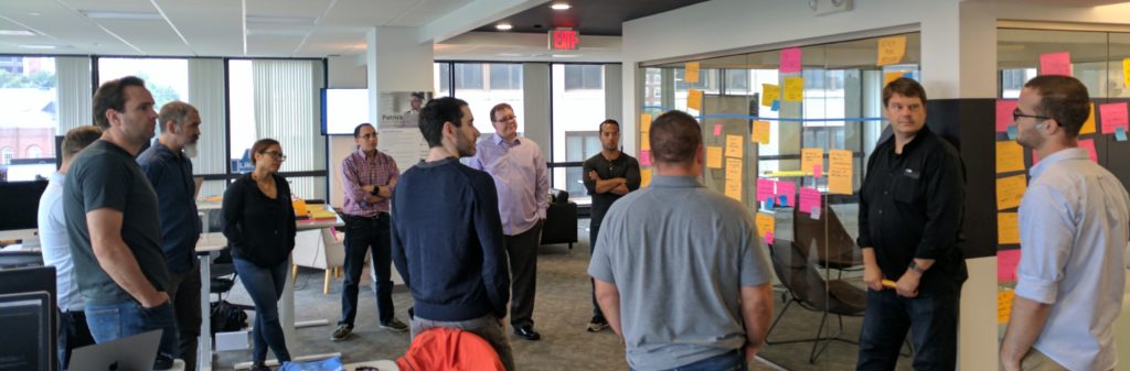 Several people standing in a circle in an office. They are all looking at one man who is speaking.